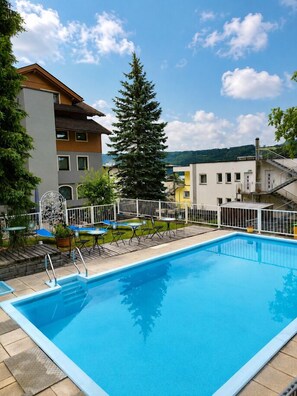 A swimming pool with a white fence, outdoor seating, and buildings in the background. - Familienhotel Steindl (Millstatt)