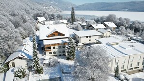 A snow-covered residential area with multiple houses, a large building with a prominent roof, and a lake in the background. - Familienhotel Steindl (Millstatt)
