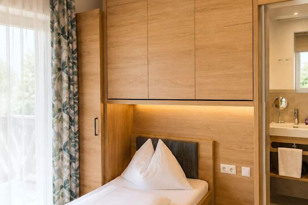 A single bed with a headboard, wooden wardrobe, and a bathroom with a mirror and sink.