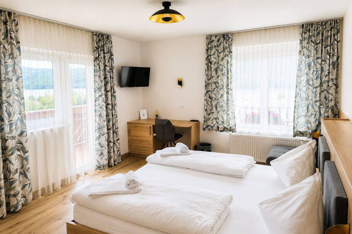A hotel room with two beds, a desk, a chair, a TV, and large windows with curtains.