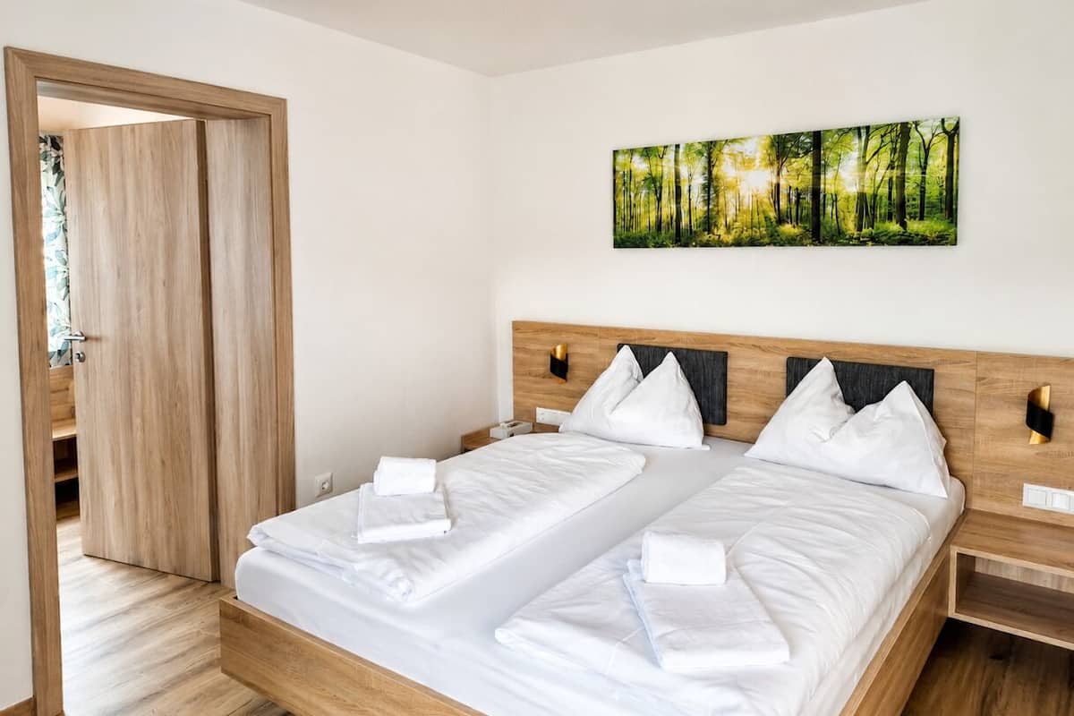 A double bed with white linens, a wooden headboard, and a forest-themed wall art.
