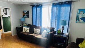 Ein Wohnzimmer mit einem dunkelbraunen Sofa, zwei Beistelltischen, einem grünen Schrank und einem Fenster mit blauen Vorhängen.