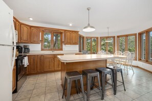 A modern kitchen with wooden cabinets, a large island with stools, and large windows offering views of trees. - Beavershore | Easy Access to Beaver Lake. Sleeps12 (Eureka Springs)