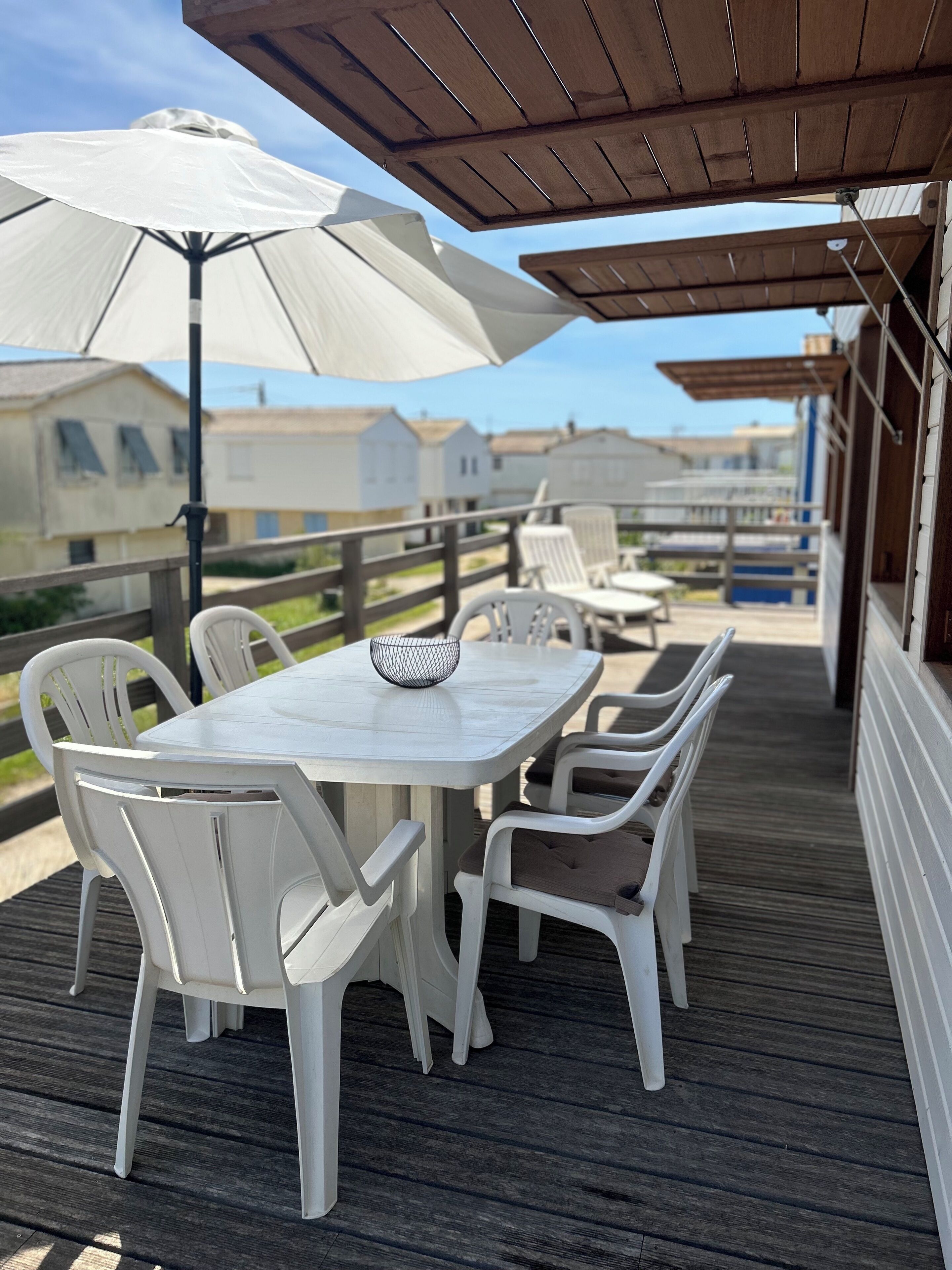 Un espace extérieur abrité, avec une table et des chaises blanches, des parasols et une vue sur des maisons en arrière-plan.