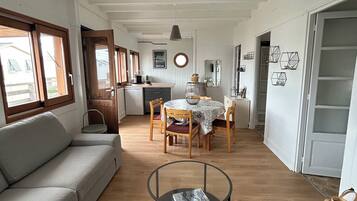 Un vaste espace de vie comprenant un canapé, une table basse ronde en verre et une table à manger avec des chaises. On aperçoit une cuisine au fond, et l’ensemble est doté de planchers en bois.