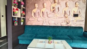 Um hall com sofá azul, mesa de centro de vidro, mural de Buda e prateleira com flores.