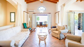 A spacious living area with a high ceiling, wooden beams, a large window, and a balcony with outdoor seating. - Villa Sea Pines in Nea Dimmata (Nea Dimmata)