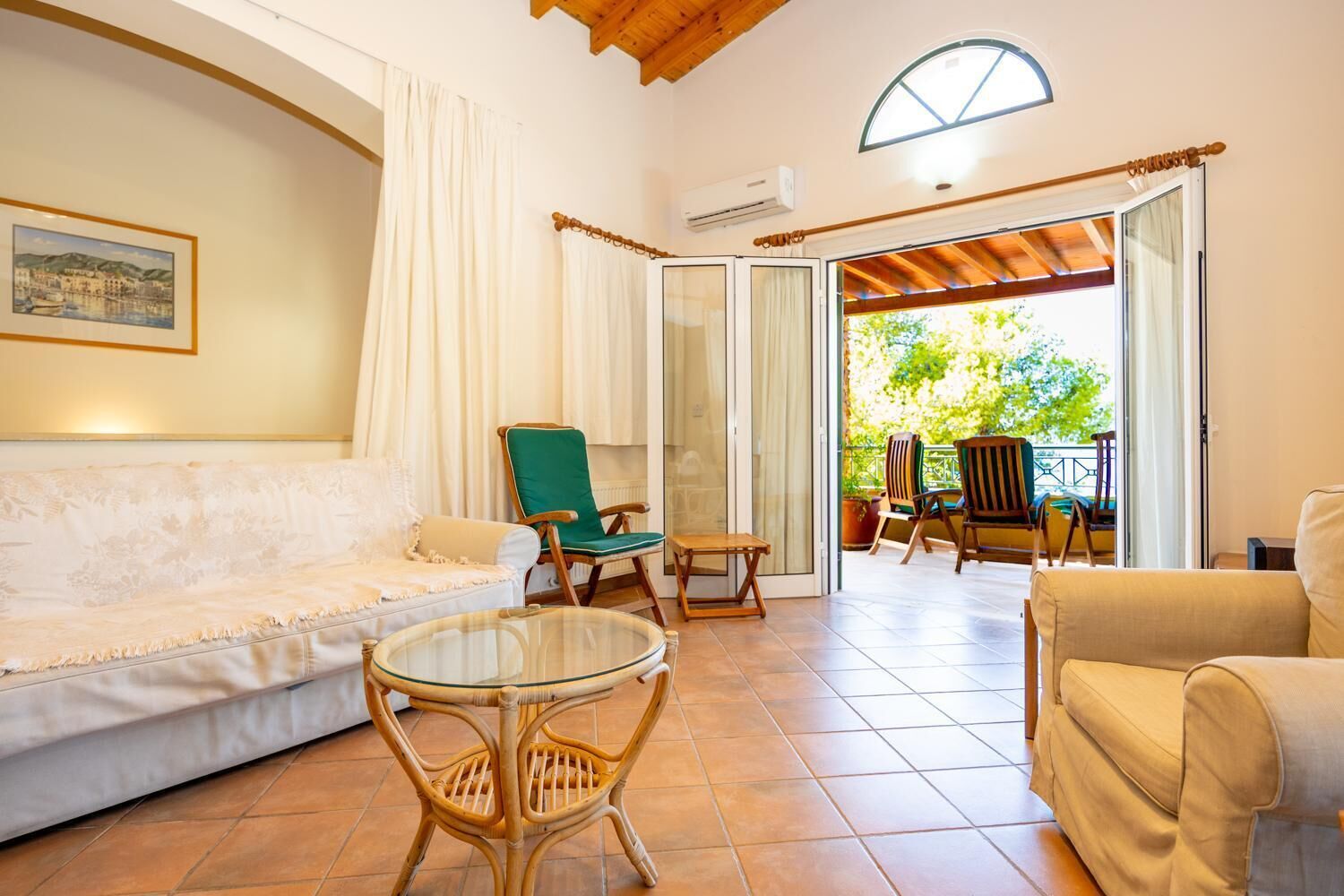 Un salon confortable avec un canapé, des fauteuils et une table en verre. Une porte-fenêtre coulissante donne accès à un patio extérieur aménagé avec des chaises et offrant une vue sur la verdure.