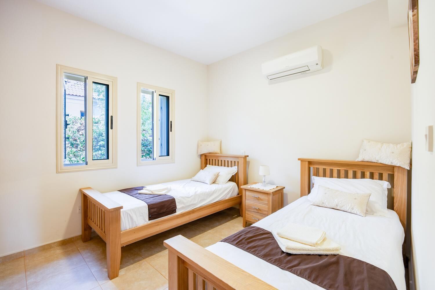 Quarto com duas camas de solteiro, uma mesa de cabeceira, uma janela e aparelho de ar condicionado.