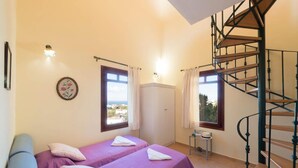 A room with two beds, a spiral staircase, a view of the sea, and a small table with a hat and a bottle. - Odysseus Villa in Plaka (Apokoronas)