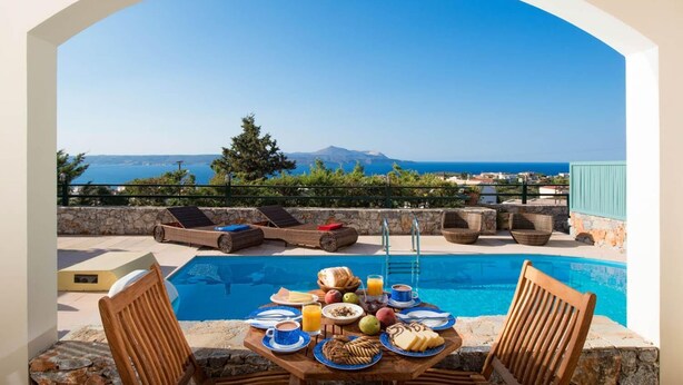 A poolside breakfast setup with a table, chairs, and a view of the sea and mountains. - Odysseus Villa in Plaka (Apokoronas)