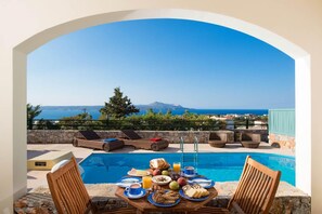 Un coin petit-déjeuner aménagé au bord de la piscine, avec une table, des chaises et une vue sur la mer et les montagnes.