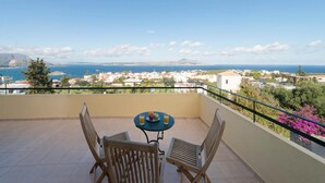 A balcony with a table and chairs, overlooking a coastal town with buildings and a mountain in the distance. - Villa Aphrodite in Plaka (Apokoronas)