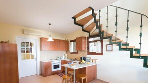 A kitchen with wooden cabinets, a white countertop, a built-in oven, a dining area with stools, and a staircase with green railings. - Villa Aphrodite in Plaka (Apokoronas)