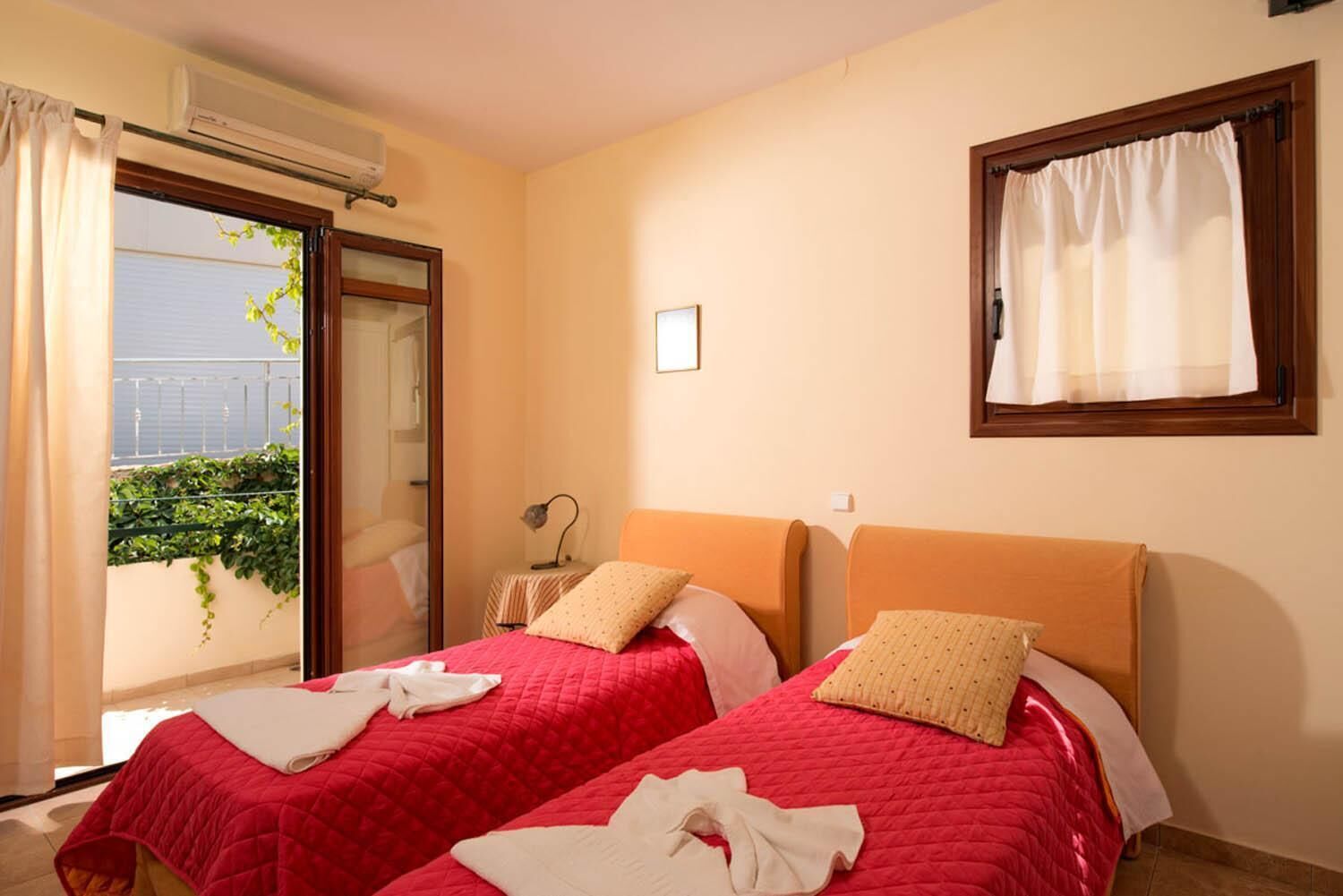 A hotel room with two beds, a window with curtains, an air conditioning unit, and a view of greenery outside.