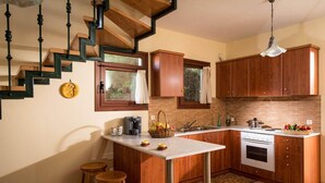 A modern kitchen with a central island, wooden cabinets, a built-in stove, and a staircase with exposed beams. - Ariadne Villa in Plaka (Apokoronas)