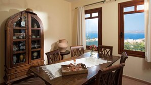 A dining area with a wooden table, chairs, a cabinet with glass doors, a vase with flowers, and a view of the sea through windows. - Ariadne Villa in Plaka (Apokoronas)