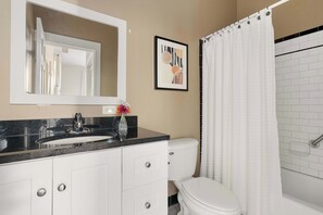 A bathroom with a white toilet, a sink with a marble countertop, a mirror above the sink, a framed picture on the wall, and a shower curtain.