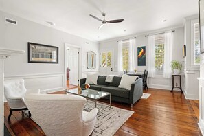 A spacious living room with hardwood flooring, a gray sofa, a white armchair, and a glass coffee table. - The Parlor | 1BD w/ Huge Porch, King Bed + Parking (Charleston)