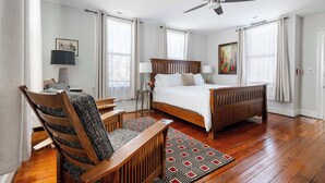 Un dormitorio con una cama de madera, dos mesitas de noche, una silla, un ventilador de techo y un tapete estampado.