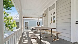 A covered porch with wooden benches and a table, white railings, and a door with a window. - The Retreat | Downtown King 2BD w/ Porch + Parking (Charleston)