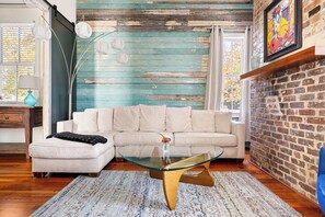 A living room with a sectional sofa, a glass coffee table, a patterned rug, a lamp, and a brick wall. - Historical 2bd Home w. Outdoor Space + Parking (Charleston)