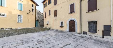 A courtyard with stone paving, buildings with arched doorways, and windows with shutters.