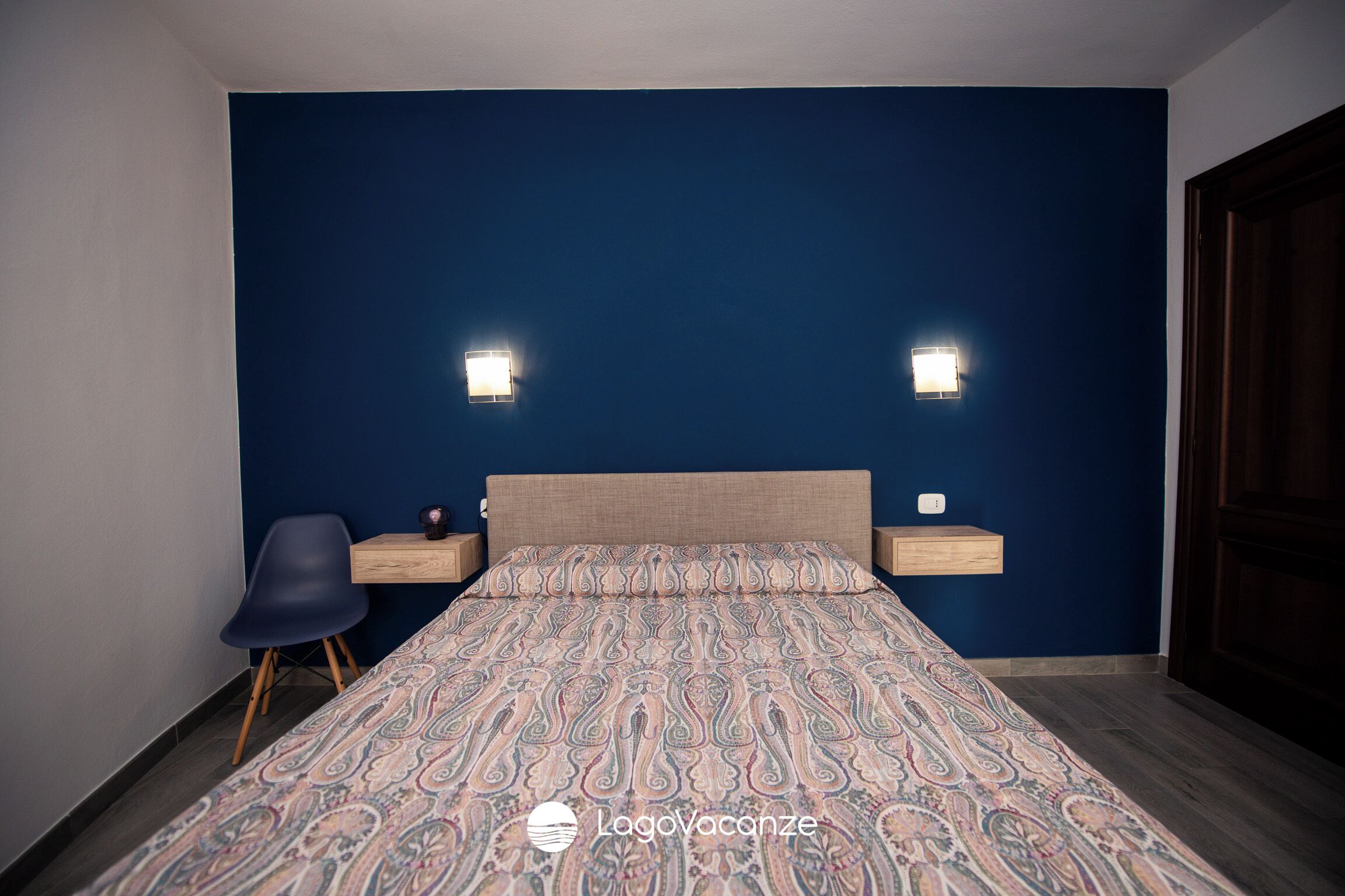 Un dormitorio con una pared azul decorativa, una colcha estampada, dos mesitas de noche con lámparas y una silla.