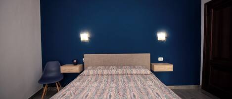 Quarto com parede azul, colcha estampada, duas mesas de cabeceira com luminárias e uma cadeira.