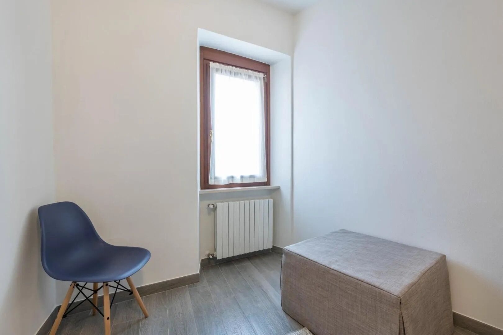 A small, clean room with a window, a radiator, a blue chair, and a small ottoman.
