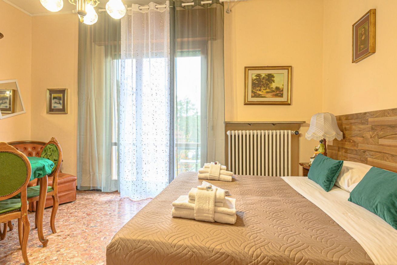 A hotel room with a bed, a chair, a radiator, a window with curtains, and a painting on the wall.