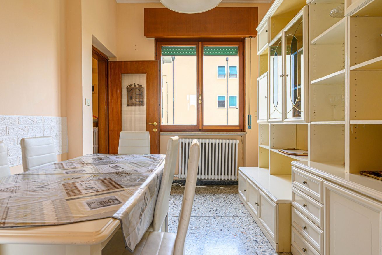 A room with a dining table, a radiator, a window with a view of buildings, and a cabinet with shelves.
