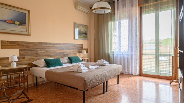 A hotel room with a bed, a rocking chair, a desk with a lamp, a window with curtains, and artwork on the walls. - Attico Vintage Colette by Rentbeat (Verona)