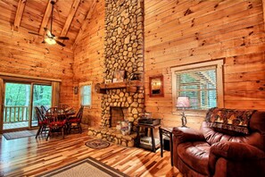 A log cabin interior with a stone fireplace, wooden floors, a leather sofa, and a dining area. - Large Log Cabin Rental Perfect for Families in Hocking Hills, Ohio (Logan)