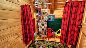 Ein Zimmer mit Holzwänden, einem roten Vorhang, einem kleinen roten Sessel und einem gelben Stuhl, sowie einem Regal mit diversen Gegenständen.