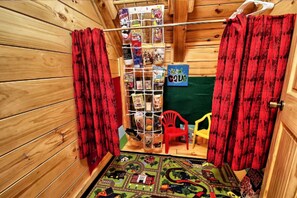 Quarto com paredes de madeira, uma cortina vermelha, uma cadeira vermelha pequena, uma cadeira amarela e uma estante com diversos objetos.