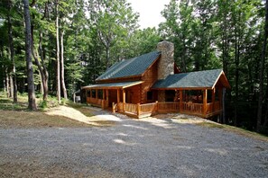 A log cabin with a stone chimney, wooden porch, and a gravel driveway. - Large Log Cabin Rental Perfect for Families in Hocking Hills, Ohio (Logan)