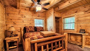 Ein Zimmer in einer Holzhütte mit Bett, Deckenventilator, Nachttisch und Fenster.