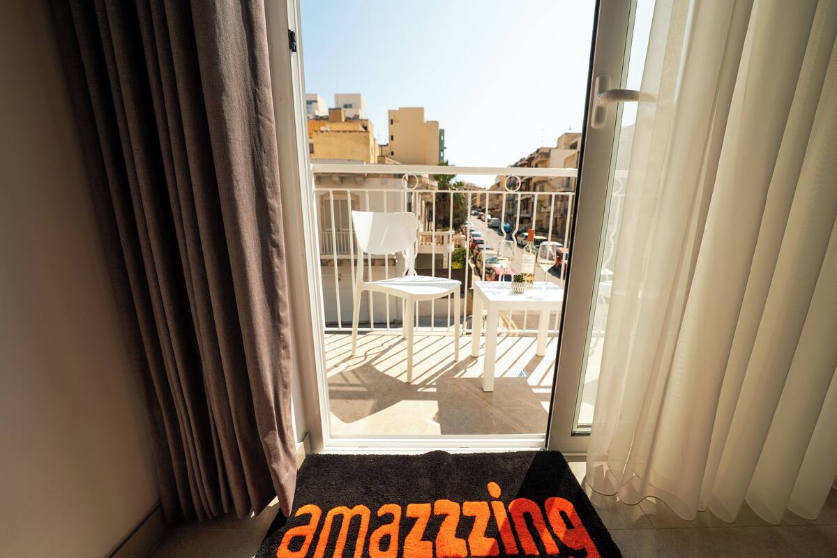 A balcony with a white chair and table, a view of buildings, and a door with curtains.