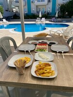 Una mesa de comedor con platos de comida, una piscina y sillones de descanso al fondo.