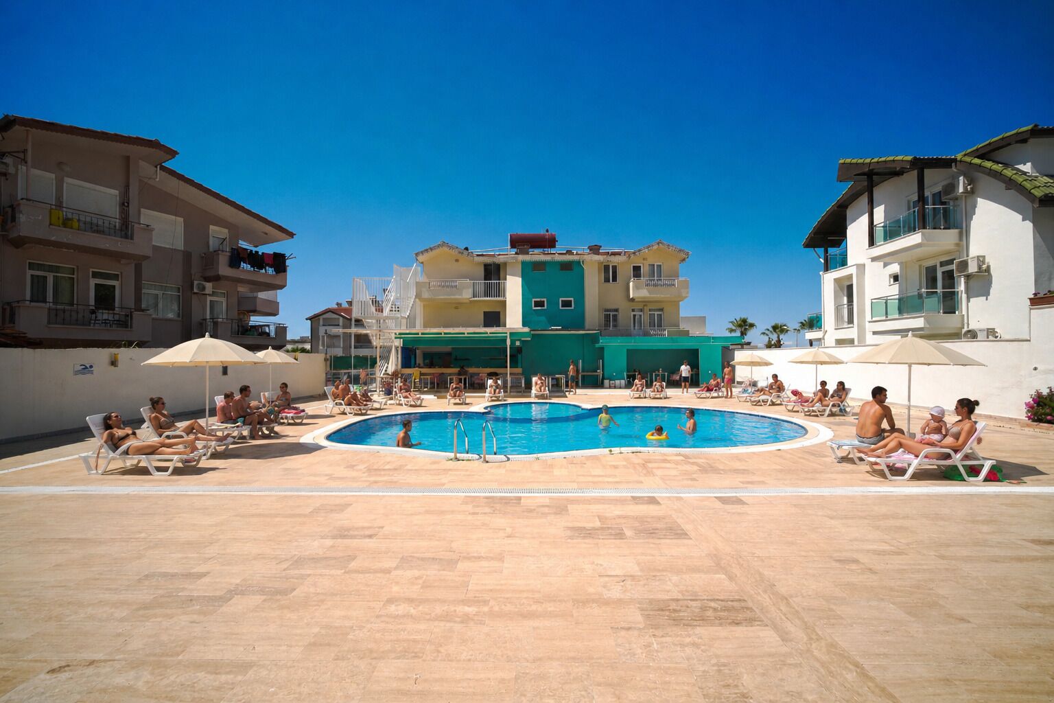 Zona de piscina con sillones de playa y sombrillas, rodeada de edificios de varios pisos.