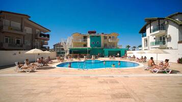 Zona de piscina con sillones de playa y sombrillas, rodeada de edificios de varios pisos.