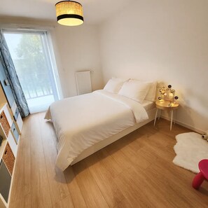Ein Schlafzimmer mit einem Bett, einem kleinen Nachttisch, einem Heizkörper, einem Fenster mit Vorhängen und einem Holzboden.