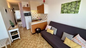 Een compacte leefruimte met een kitchenette, een bank met gestreepte kussens, een kleine tafel met een potplant en een decoratief wandobject.