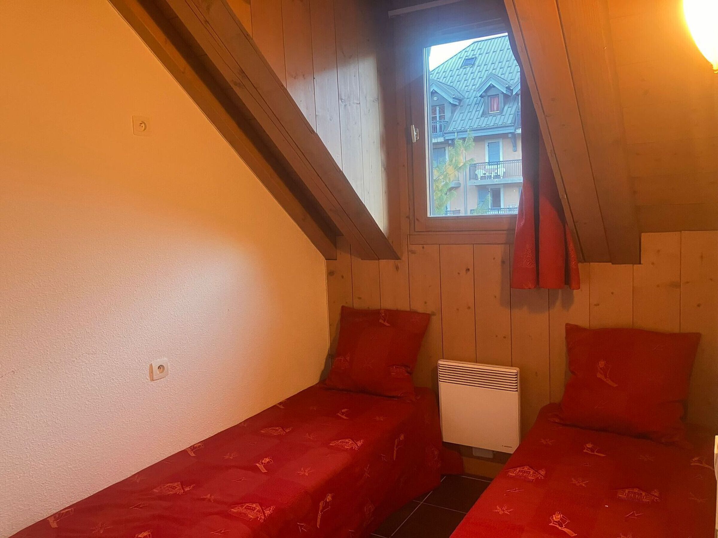 A small, cozy room with wooden walls and a sloped ceiling. There are two beds with red bedding, a window with a view of a house, and a white heater.