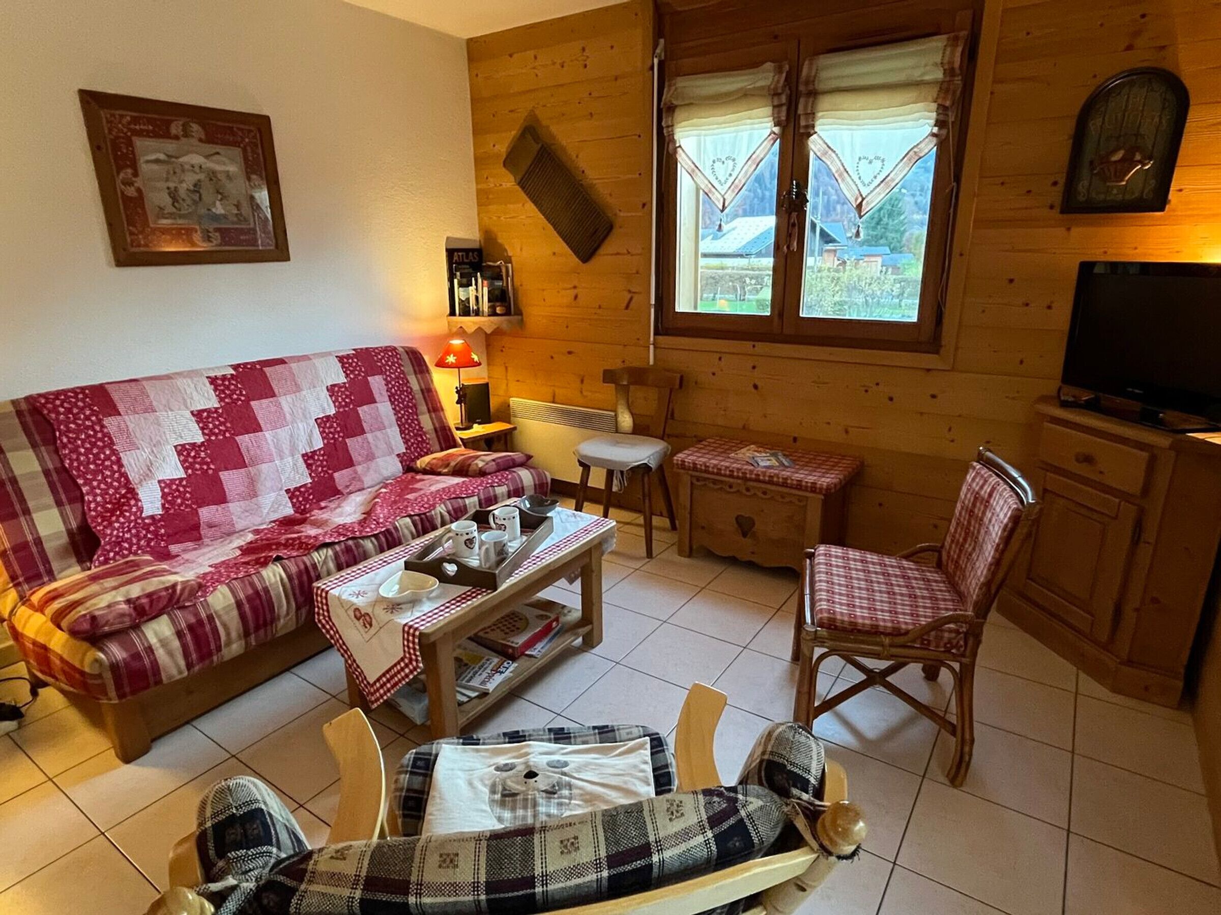 Un interior de cabaña acogedor con piso de madera, un sofá a cuadros rojo y blanco, un sillón escocés, una mesita con una bandeja de tazas y un televisor sobre un mueble de madera.