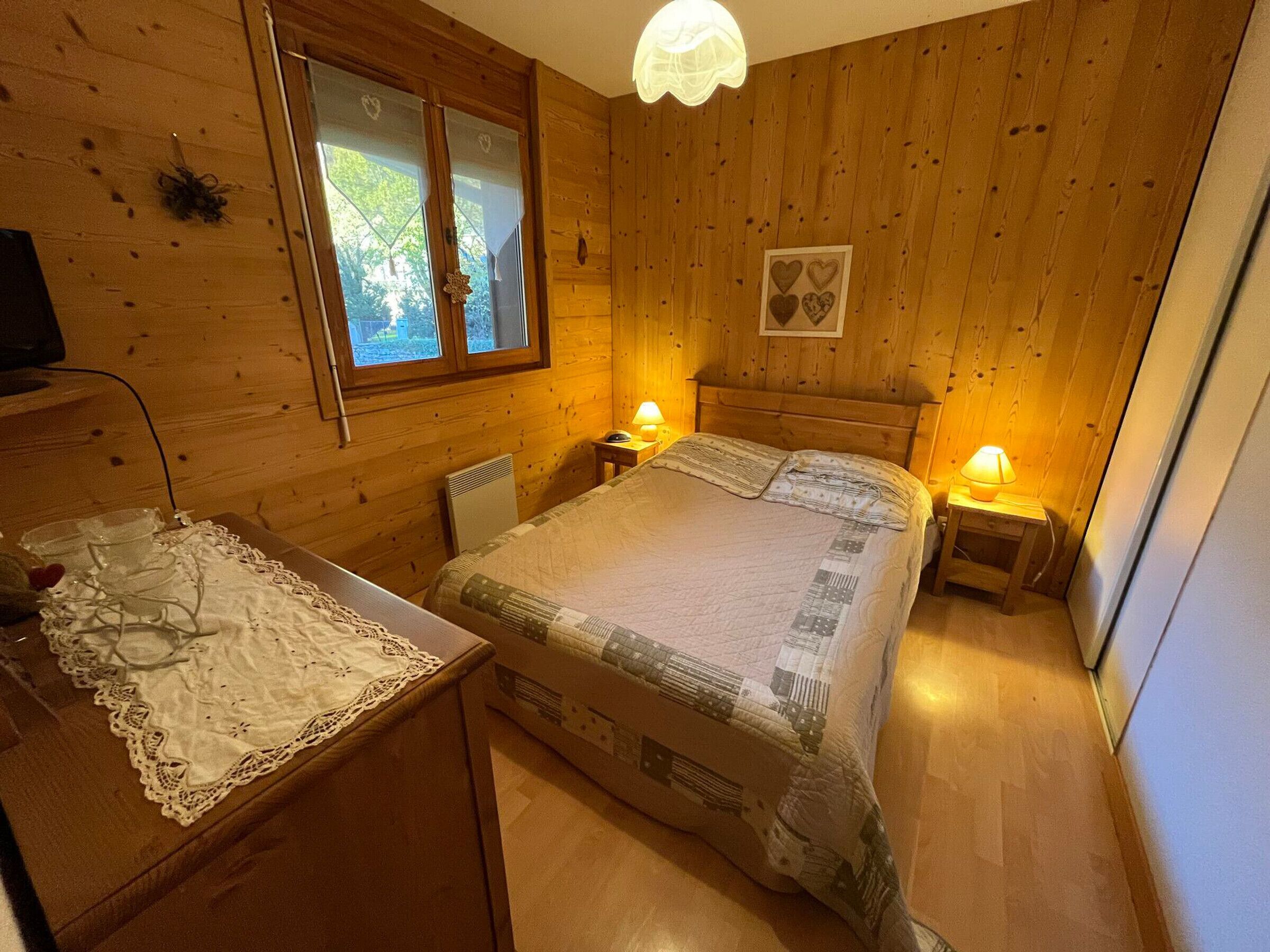 A cozy wooden room with a bed, bedside lamps, a desk with a lace tablecloth, and a window with heart-shaped curtains.