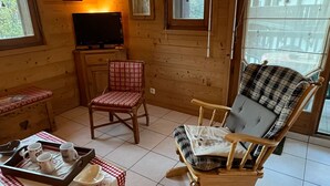 Interior acogedor de una cabaña con paredes de madera, un televisor, un sillón mecedor, una mesa de comedor con una bandeja de tazas y una ventana con vistas a un edificio.