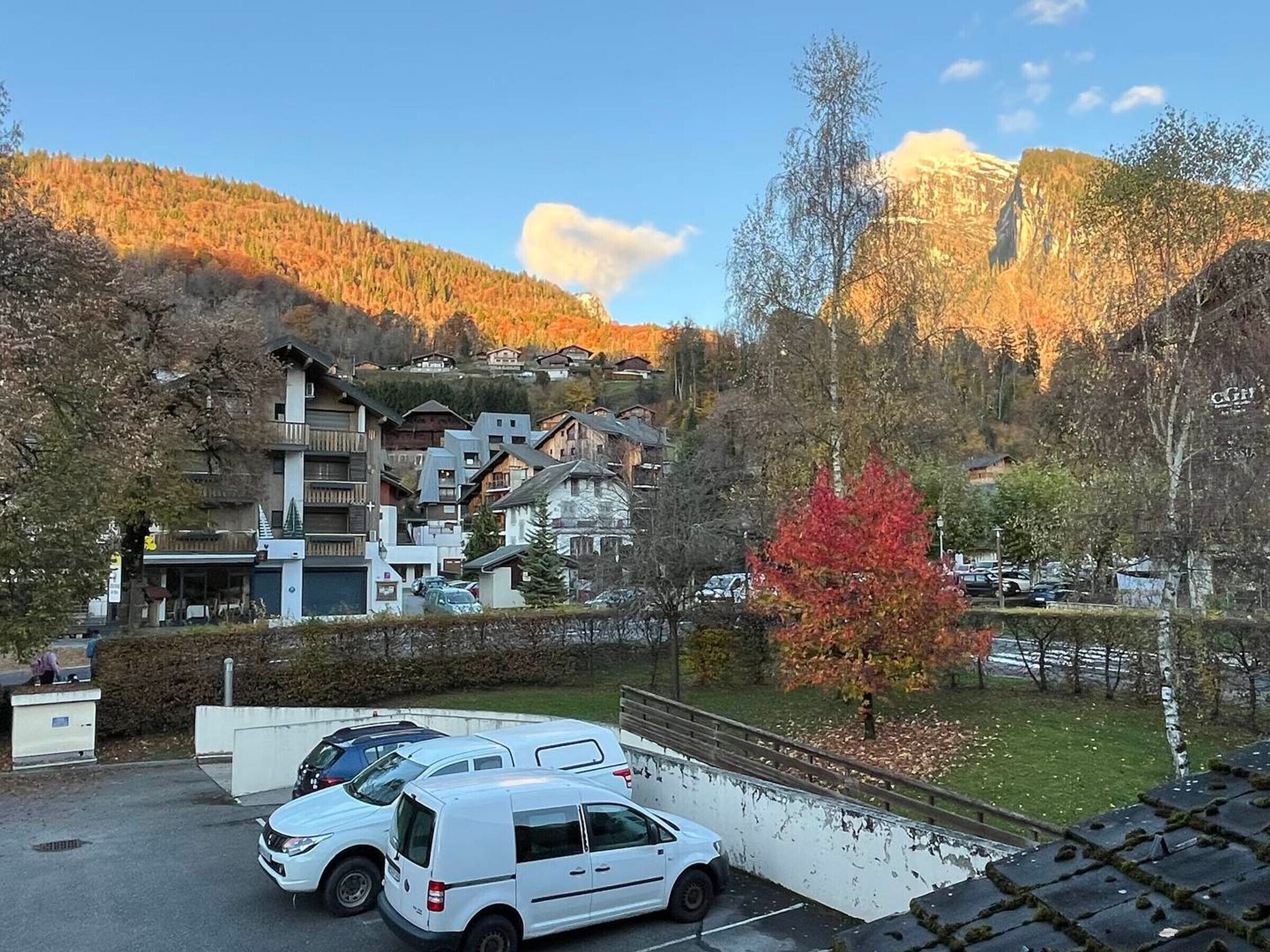 A parking area with a white van and several cars, surrounded by buildings with balconies and a mountain backdrop.