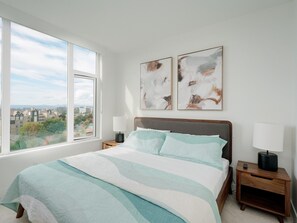 A bedroom with a large window, a bed with a striped bedspread, two bedside tables with lamps, and abstract art on the wall. - Royal Vista - Exceptional Views, AC and lots of Amenities by Victoria Prime! (Victoria)